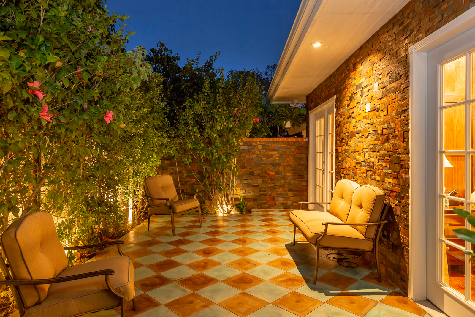 Outdoor patio area at dusk with cushioned chairs and a loveseat arranged on a checkered tile floor. The patio is bordered by a stone wall and lush green bushes with pink flowers, illuminated by warm lighting.