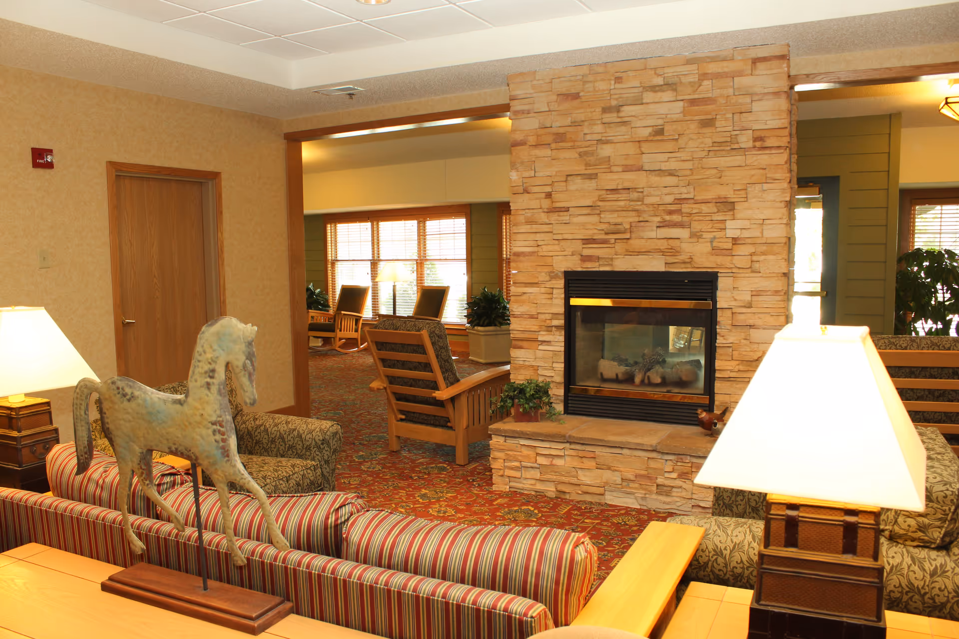Interior view of a senior living facility lounge area featuring a stone fireplace, patterned carpet, wooden rocking chairs near large windows, a striped sofa with a decorative horse sculpture on the back, and table lamps providing warm lighting.