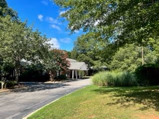 A driveway leading to a single-story building surrounded by green trees and bushes under a blue sky with some clouds.