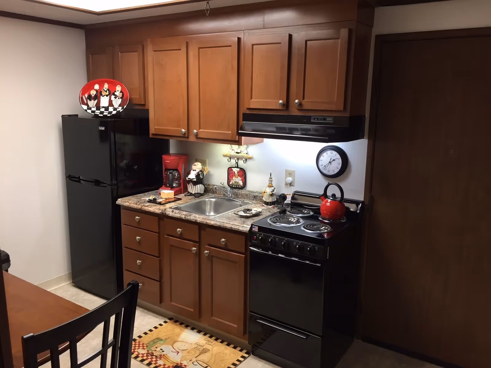 A compact kitchen area with wooden cabinets, a black refrigerator, a black stove with a red kettle on top, a stainless steel sink, and a countertop with a red coffee maker and decorative items. A wall clock is mounted above the stove, and a decorative plate is displayed on top of the refrigerator.