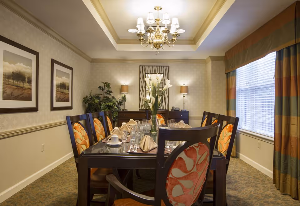 A formal dining room with a rectangular dark wood table set for eight people. The chairs have orange patterned upholstery. The table is set with glasses, napkins, and a vase with white flowers in the center. The room has beige walls with two framed landscape paintings, a large window with striped curtains, a chandelier, and two wall lamps above a sideboard with a decorative mirror and plants.