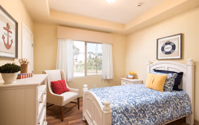A bright and cozy bedroom with a white wooden bed featuring blue and white floral bedding and yellow accent pillows. There is a white nightstand with a small plant and a book, a white dresser with a potted plant and decorative items, and a beige armchair with a red cushion. The room has a large window with white curtains and light yellow walls adorned with nautical-themed artwork.