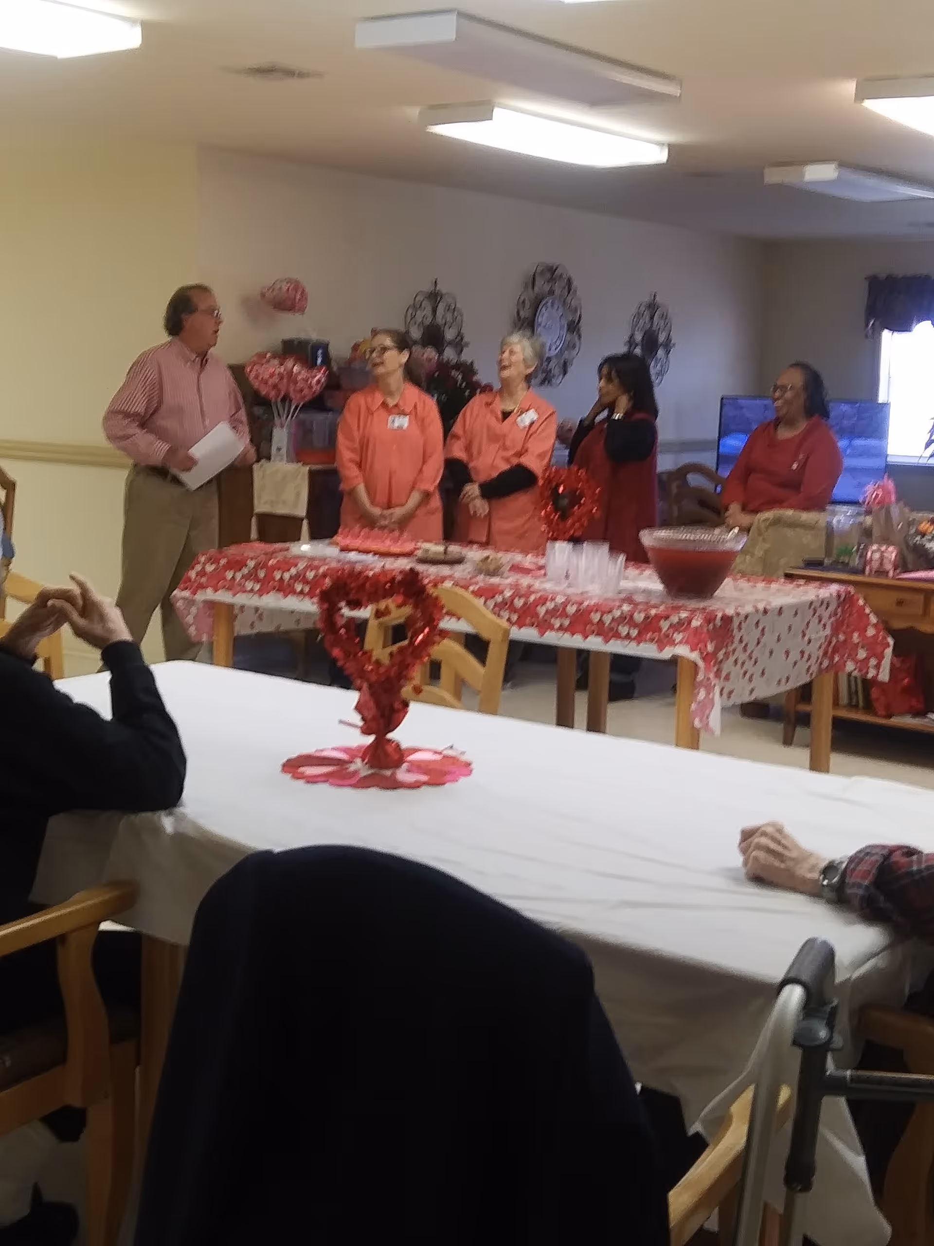 A group of five people standing behind a table decorated with a red and white tablecloth and Valentine-themed decorations in a common room. The table has a large punch bowl and cups. Two elderly people are seated at a table in the foreground, one with a walker nearby. The room has beige walls and ceiling lights.