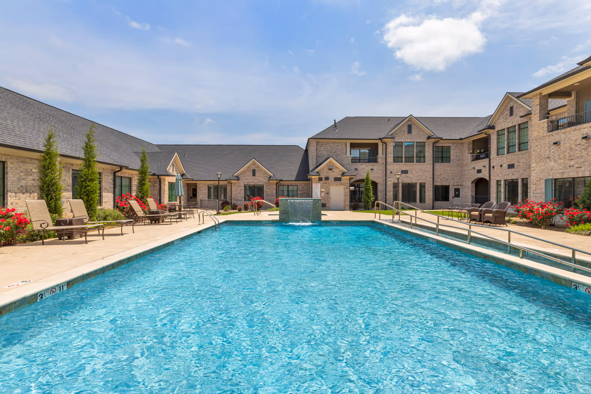 Outdoor swimming pool in the courtyard of a senior living facility with lounge chairs, umbrellas, and brick buildings surrounding the pool under a partly cloudy sky.