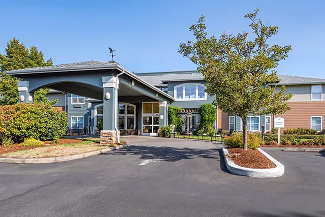 Front exterior view of Brookdale Rose Valley Scappoose senior living facility with a covered entrance, landscaped bushes and trees, and a clear blue sky.