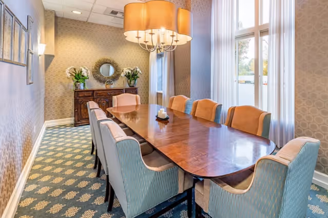 A bright private dining room with a long polished wooden table surrounded by upholstered chairs, a chandelier overhead, and a sideboard against the wall.
