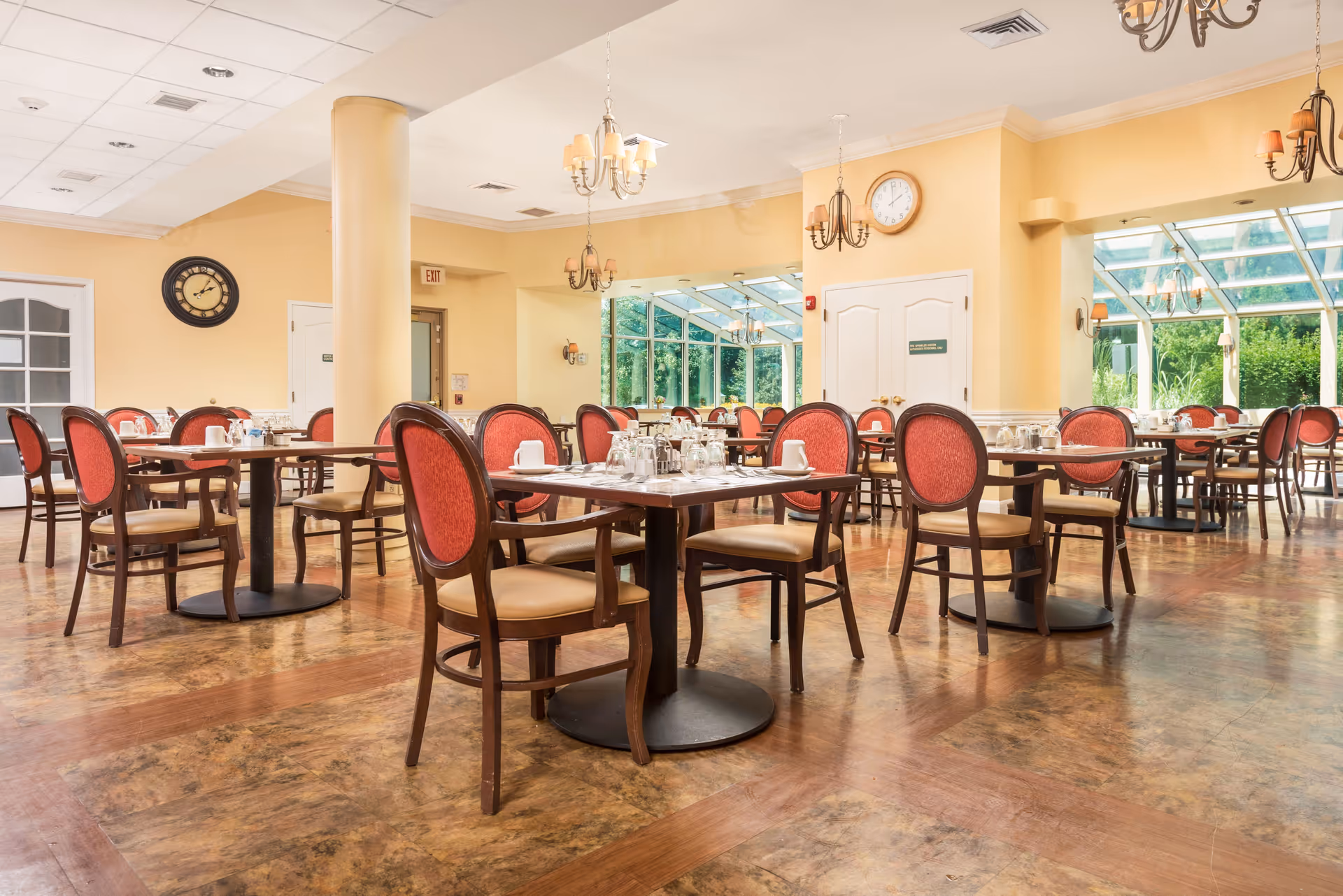 A bright dining room with multiple tables and chairs. The chairs have wooden frames with red cushioned backs and beige seats. Tables are set with cups, glasses, and condiments. The room has yellow walls, a clock on the wall, chandeliers hanging from the ceiling, and large windows letting in natural light with a view of greenery outside.