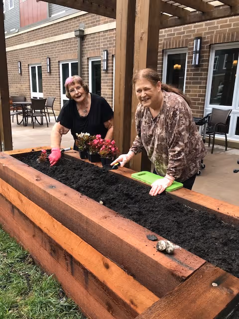 Two elderly women gardening together in a raised wooden planter box outside a brick building. One woman is wearing pink gardening gloves and holding a small plant, while the other is using a small gardening tool. There are potted flowers on the edge of the planter, and patio furniture is visible in the background.