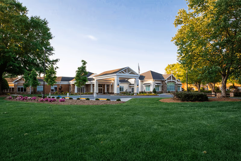 Front exterior of a senior living building with a covered entrance, wide lawn, flower beds, and trees under a clear sky.