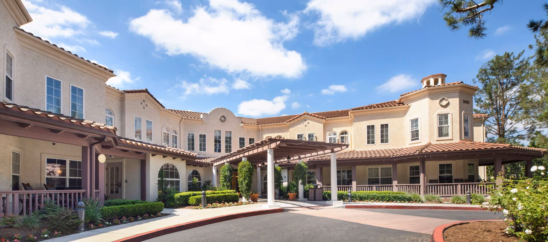 Exterior view of a senior living facility building with beige stucco walls and red tile roofing under a blue sky with scattered clouds. The entrance features a covered walkway with columns and greenery around the pathway.