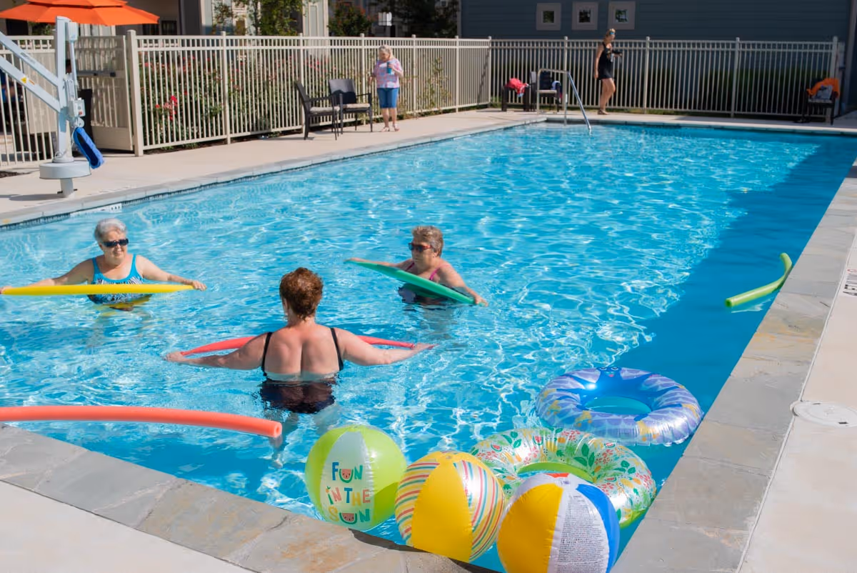 Three elderly women in swimsuits are enjoying a sunny day in an outdoor swimming pool, each holding a colorful pool noodle. Several inflatable beach balls and rings float near the pool edge. In the background, a fenced area with chairs and two other people standing near the pool is visible.