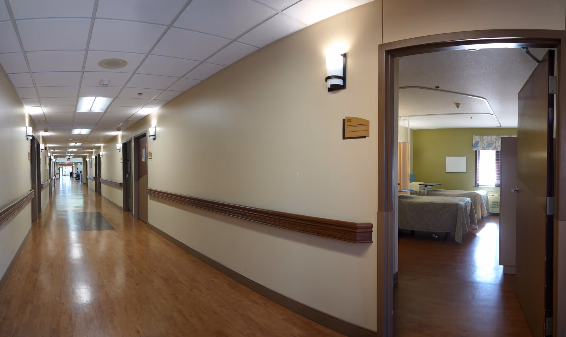 A long, well-lit hallway in a senior living facility with wooden floors and beige walls. On the right side, an open door reveals a bedroom with two beds, a window with blinds, and light green walls.