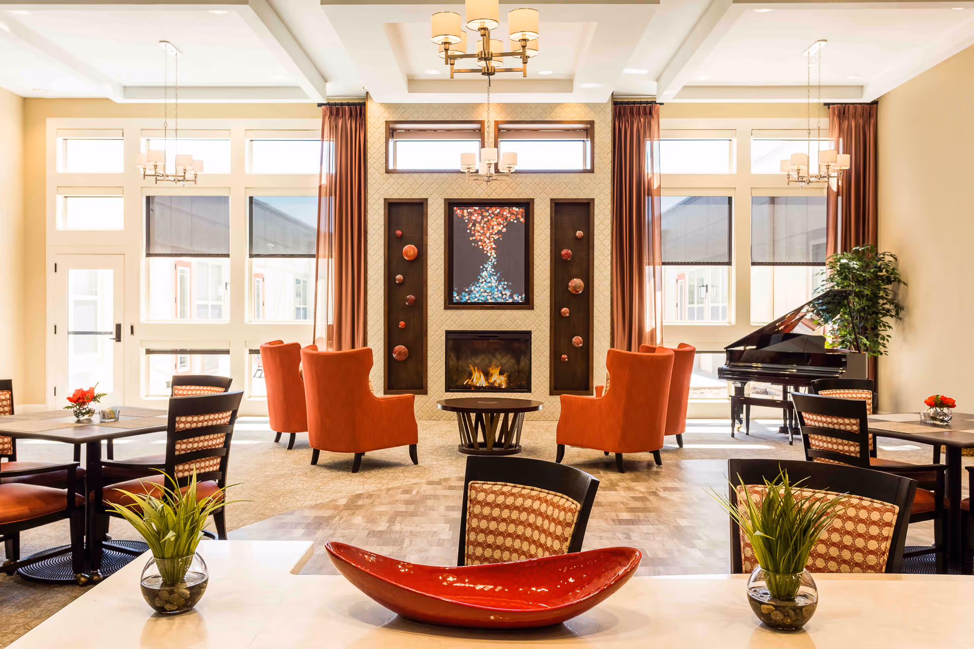 Bright communal lounge and dining area with tables, red armchairs around a fireplace, and a grand piano.