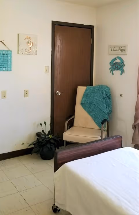 A corner of a nursing home room with a closed wooden door, a beige chair with a teal blanket draped over it, a potted plant on the floor, and part of a bed with white bedding. The walls have decorative signs including a crab and a sign that says 'Choose Happy'.