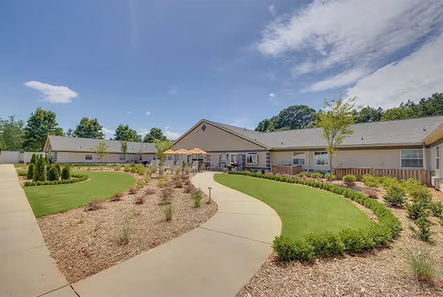 Outdoor view of a senior living facility with a curved concrete walkway surrounded by landscaped garden beds and green grass. The single-story building has a light-colored exterior with several windows and a patio area with umbrellas and seating. Trees and blue sky with some clouds are visible in the background.