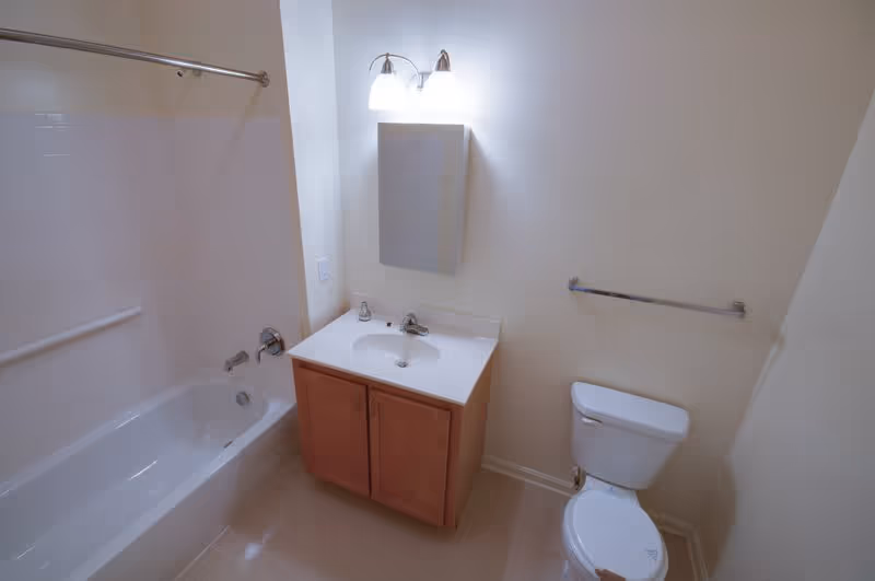 A clean bathroom featuring a white bathtub with a shower curtain rod, a wooden vanity with a white sink and faucet, a wall-mounted mirror cabinet above the sink, a toilet, and a towel bar on the wall.