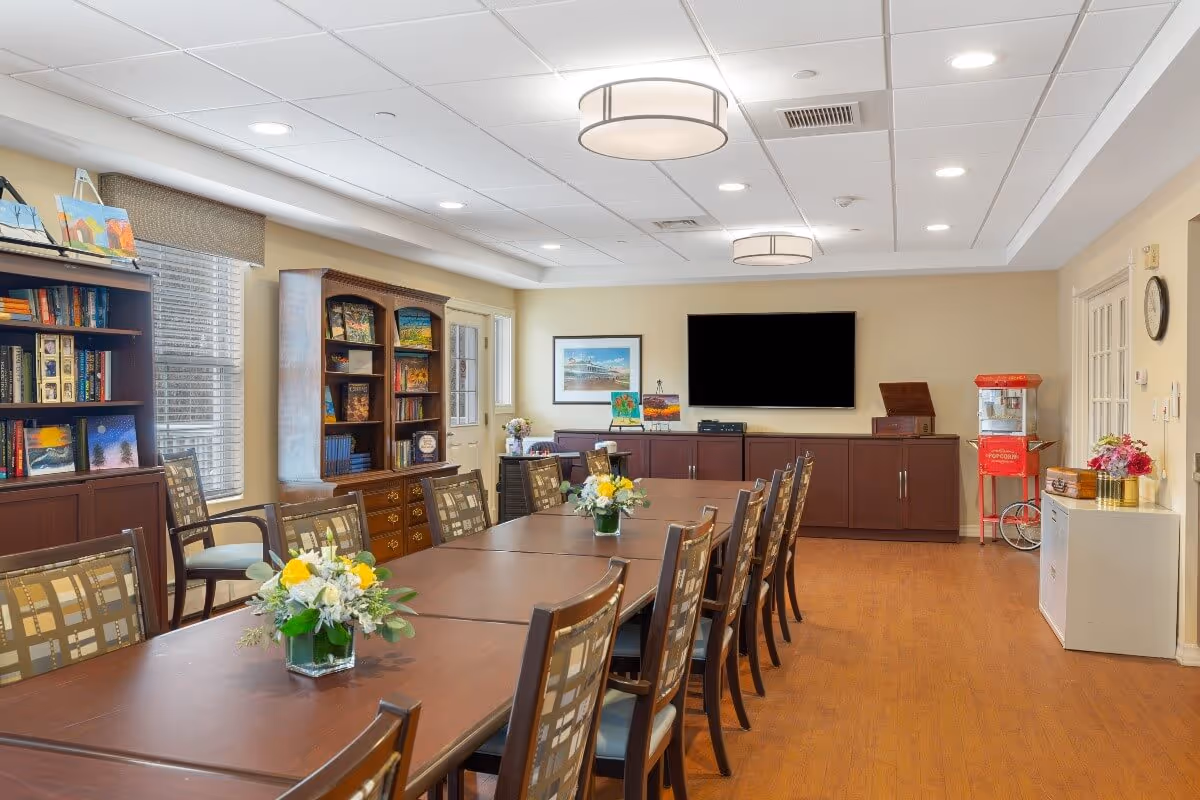 A well-lit room with a long wooden table surrounded by chairs, decorated with flower arrangements. The room features bookshelves filled with books and artwork, a large flat-screen TV mounted on the wall, a popcorn machine in the corner, and a clock on the wall. The floor is wooden, and the ceiling has recessed lighting and modern circular light fixtures.