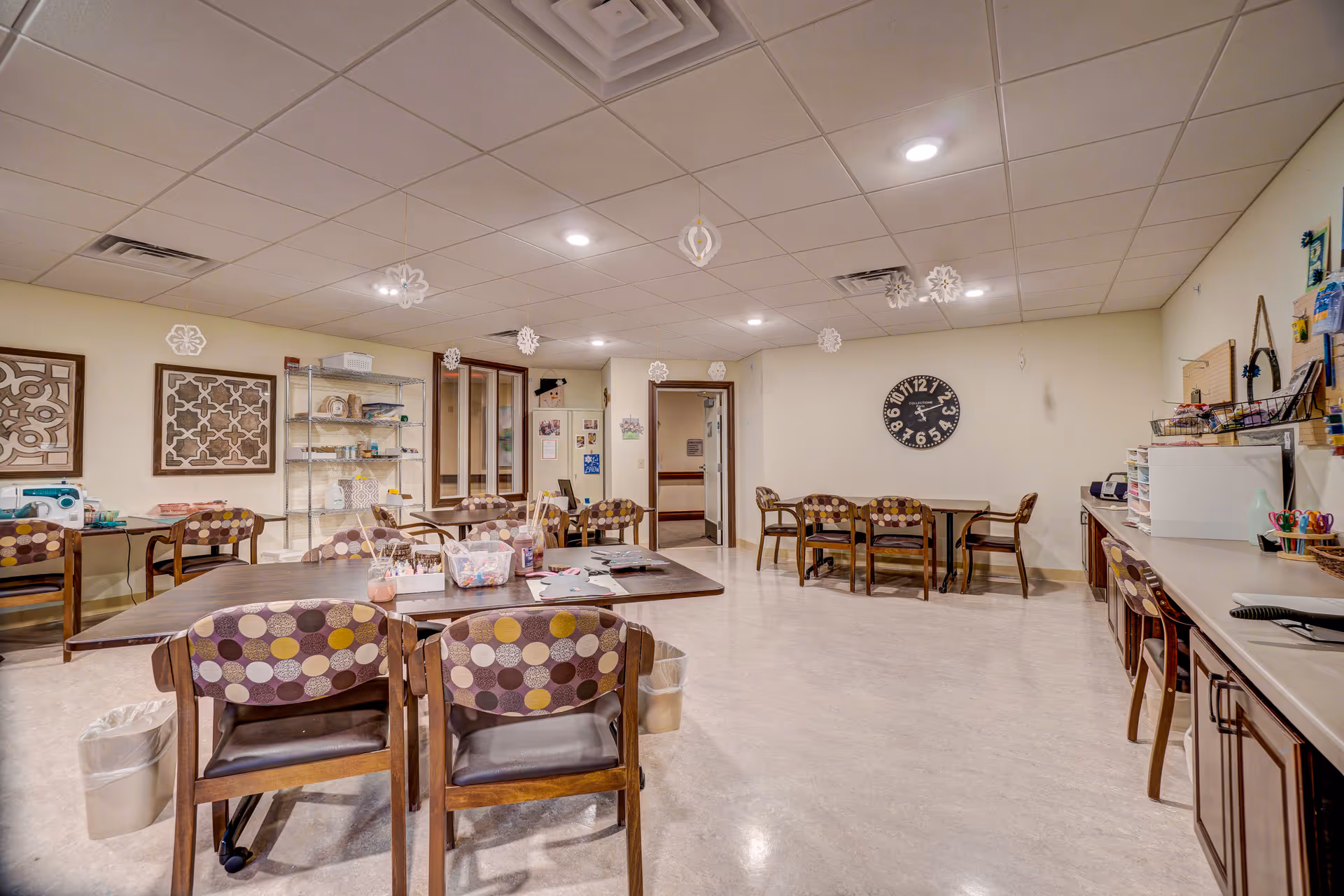 A spacious activity or craft room with several tables and chairs featuring circular patterned upholstery. The room has a light-colored floor and ceiling with recessed lighting. There are craft supplies on the tables and shelves along the walls, including a sewing machine and various containers. Decorative snowflakes hang from the ceiling, and a large clock is mounted on the far wall.