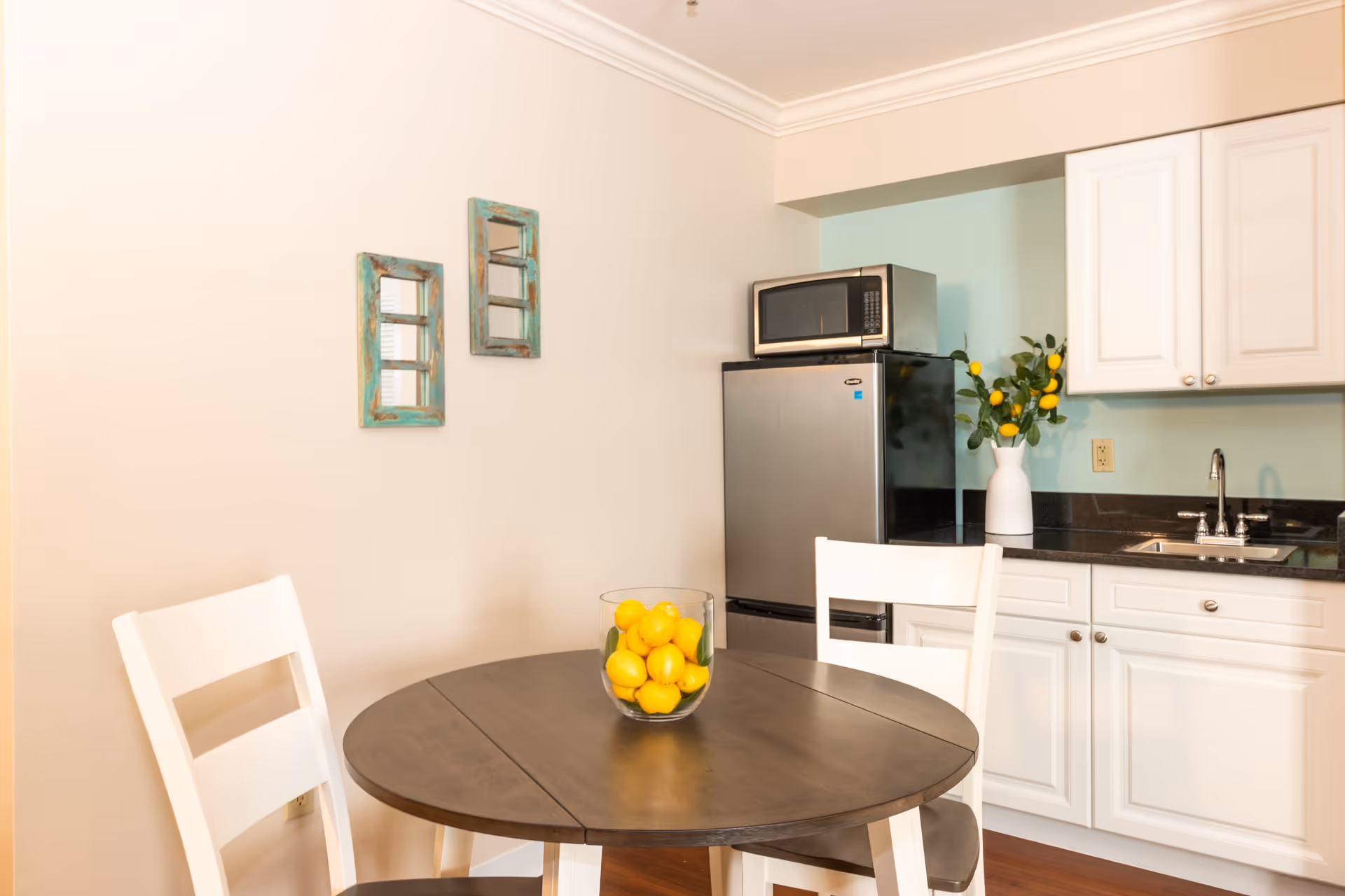A small kitchen dining area with a round wooden table and two white chairs. On the table is a glass bowl filled with lemons. In the background, there is a stainless steel refrigerator with a microwave on top, white cabinets, a sink, and a vase with lemon branches. Two decorative turquoise-framed mirrors hang on the beige wall.