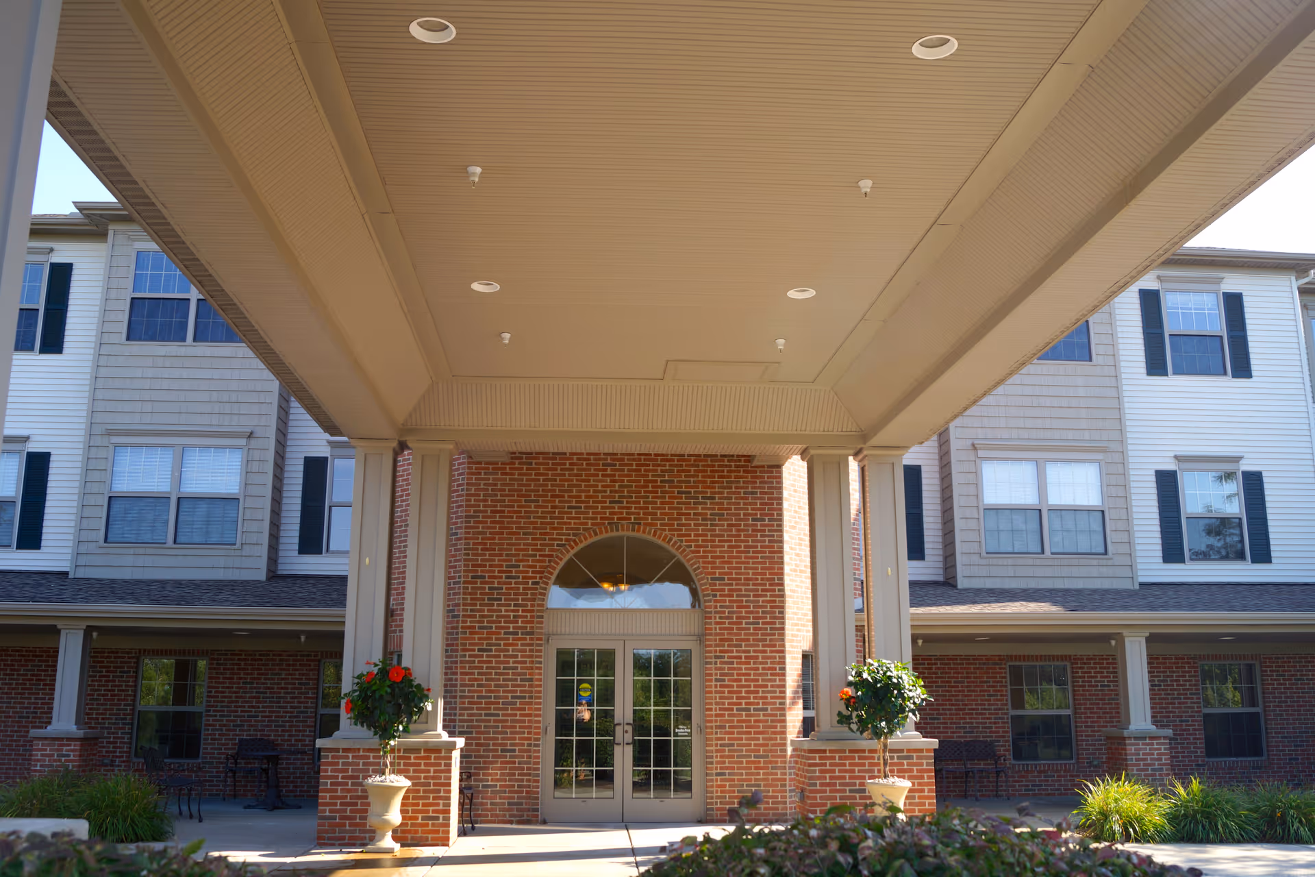 Covered main entrance of a multi-story brick and siding senior living building with double doors and potted plants.