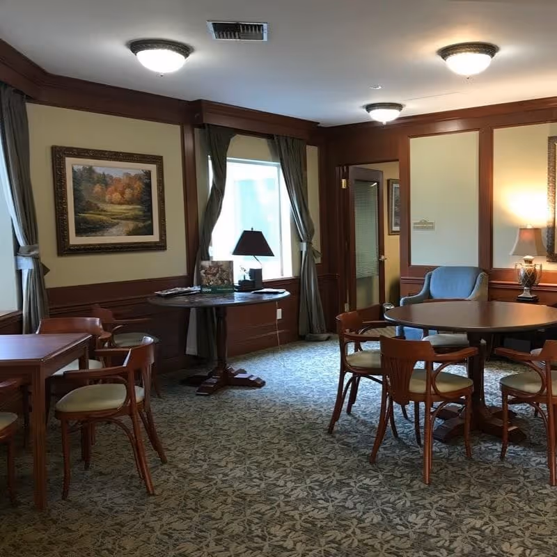 A cozy interior room with multiple wooden tables and chairs arranged on a patterned carpet. The walls have wooden paneling and are decorated with framed landscape paintings. There are windows with curtains, a table lamp on a round table, and ceiling lights providing illumination.