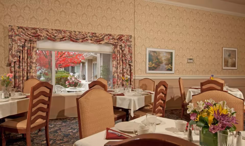 A cozy dining room with several tables covered in white tablecloths, set with cups, glasses, and napkins. Floral arrangements decorate the tables. The room features patterned wallpaper, framed landscape paintings on the walls, and a large window with floral curtains showing an outdoor garden with red-leaved trees and white patio furniture.