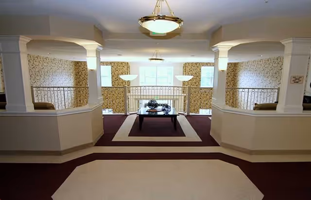 Interior view of a common area with patterned wallpaper, two large pillars with railings, a central coffee table with decorative items, and hanging ceiling lights. The area is carpeted with a maroon and beige design.