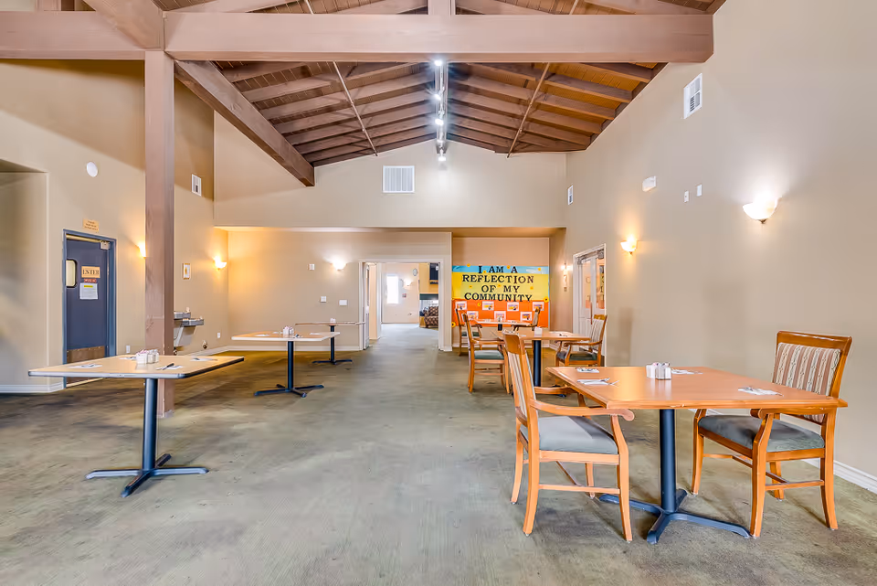 Interior view of a senior living facility dining room with several wooden tables and chairs spaced apart. The room has beige walls, a high wooden beam ceiling, and soft wall lighting. A colorful bulletin board at the far end reads 'I AM A REFLECTION OF MY COMMUNITY.'