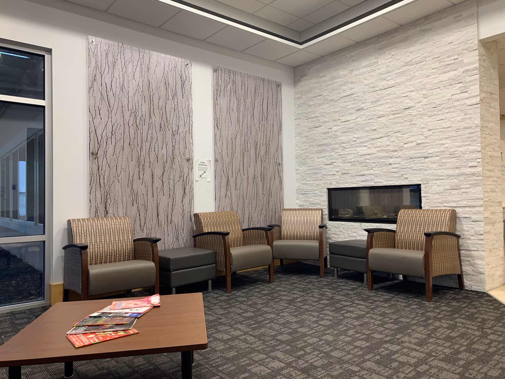A cozy seating area with four cushioned armchairs arranged around a modern rectangular coffee table with magazines on it. The room features a white textured stone wall with a built-in horizontal fireplace and two large decorative panels with a branch pattern on the adjacent wall. There is a large window on the left side, and the floor is carpeted with a patterned design.