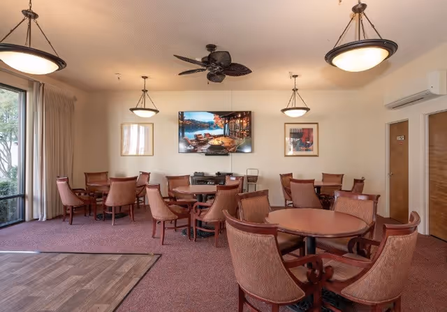 A senior living facility common area with multiple round tables and upholstered chairs arranged for group seating. The room has carpeted flooring with a small section of wood flooring, large windows with curtains, ceiling fans, and hanging light fixtures. A flat-screen TV is mounted on the wall, flanked by framed artwork.