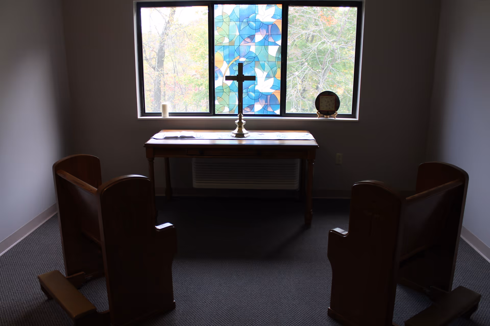 A small chapel room with two wooden pews facing a wooden table. On the table is a brass cross, an open book, a candle, and a decorative plate. Behind the table is a window with a stained glass panel featuring blue, green, and orange colors and white doves, flanked by clear glass panes showing trees outside.