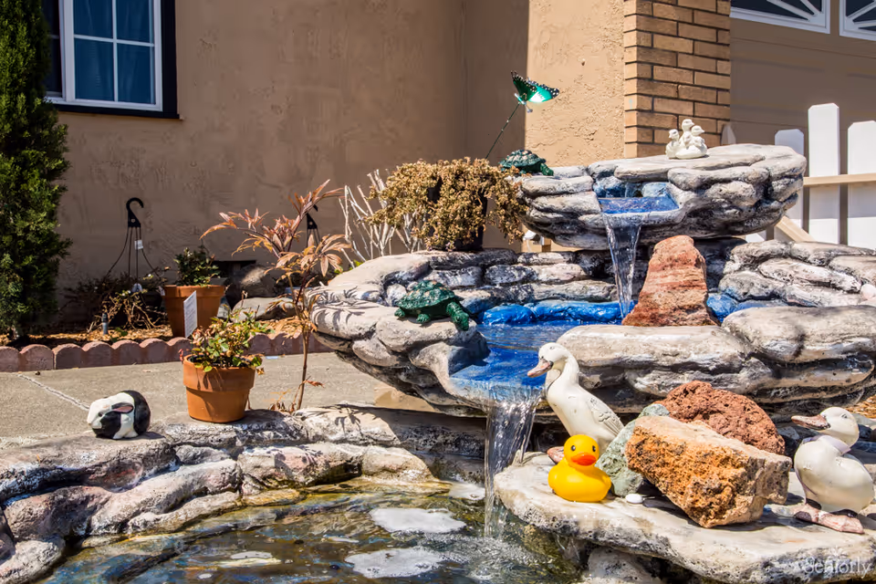 An outdoor tiered stone fountain with flowing water, decorative duck statues and a rubber duck in front of a house.