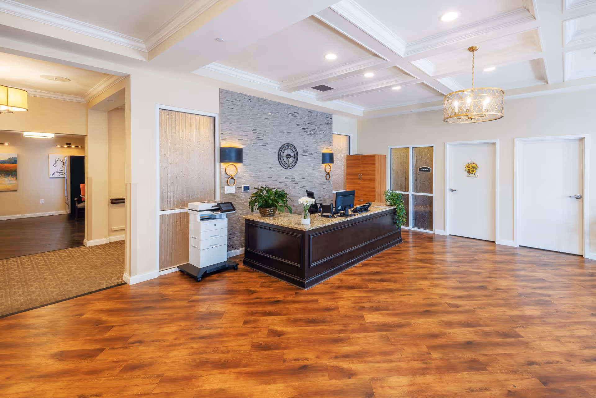 Bright reception lobby with a dark wood front desk, wood-look flooring, decorative lighting, and several doors along the back wall.