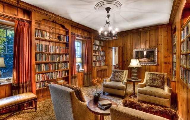 A cozy library room with wood-paneled walls and built-in bookshelves filled with books. The room features three upholstered armchairs arranged around a small round wooden table with a book and a small plant on it. There are two windows with red curtains, a bench beneath one window, a standing lamp, and a chandelier hanging from the ceiling. A framed picture hangs on the wall.