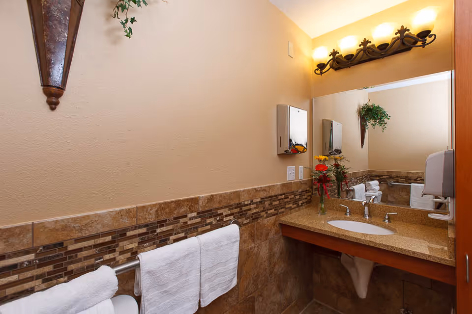 Tidy tiled bathroom with a granite sink countertop, mirror and lighting, towel rack with white towels, and decorative flowers and wall accents.