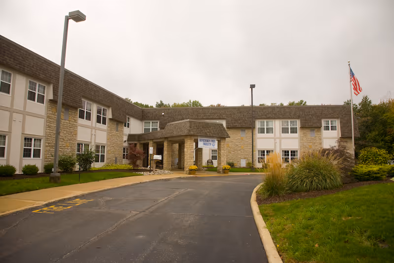 Front entrance of a two-story senior living facility with a driveway, flagpole, and landscaped grounds.