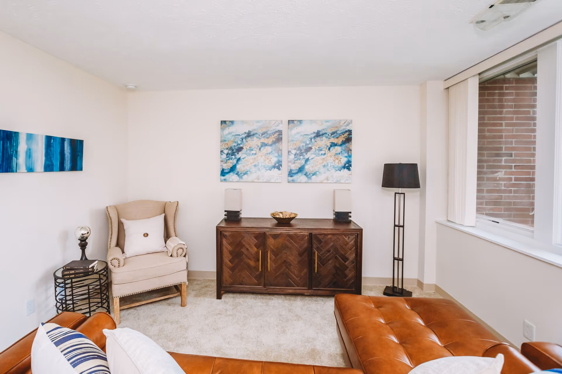 A cozy living room with a brown leather sectional sofa adorned with white and blue pillows. There is a beige armchair with a white pillow and a small round black side table holding books and a decorative item. Against the wall is a dark wooden cabinet with two lamps and a decorative bowl on top. Above the cabinet are two abstract blue and beige paintings. A tall black floor lamp stands near a large window with vertical blinds, showing a brick exterior wall outside.