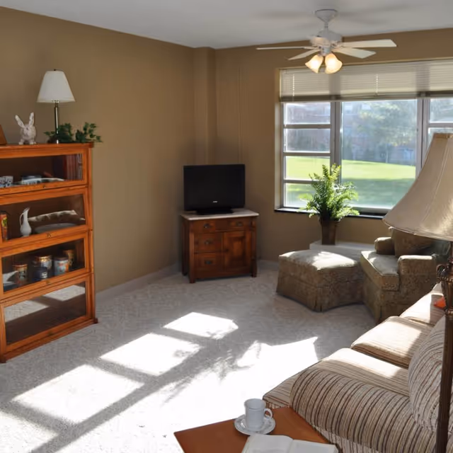 A cozy living room with beige walls and carpeted floor. The room features a striped sofa, a wooden coffee table with a cup and saucer and an open book, a wooden cabinet with glass doors displaying various items, a small wooden TV stand with a flat-screen TV, a cushioned armchair with an ottoman near a large window letting in natural light, and a ceiling fan with lights.