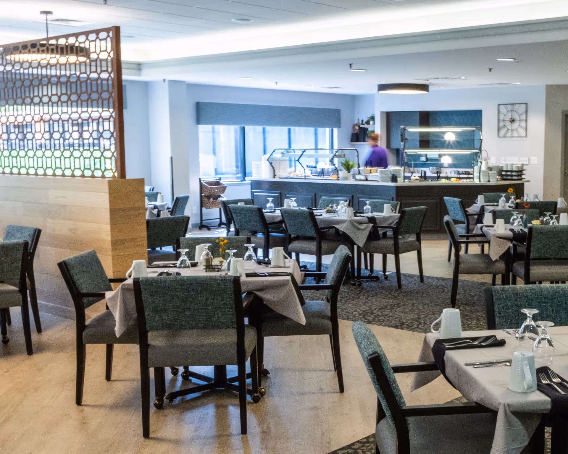 A dining room in a senior living facility with multiple tables set with white tablecloths, napkins, glasses, and mugs. Chairs with teal upholstery surround the tables. In the background, there is a buffet area with food warmers and a person in a purple shirt. The room has large windows and modern decor.
