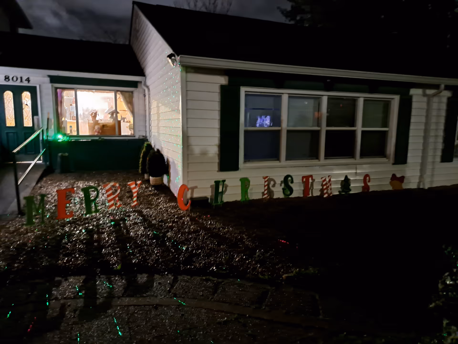 Night view of the front exterior of a residential care facility with white siding and green shutters. The house number 8014 is visible near the green front door. A festive 'MERRY CHRISTMAS' sign is displayed on the ground in front of the building, with some holiday decorations visible inside through the window.