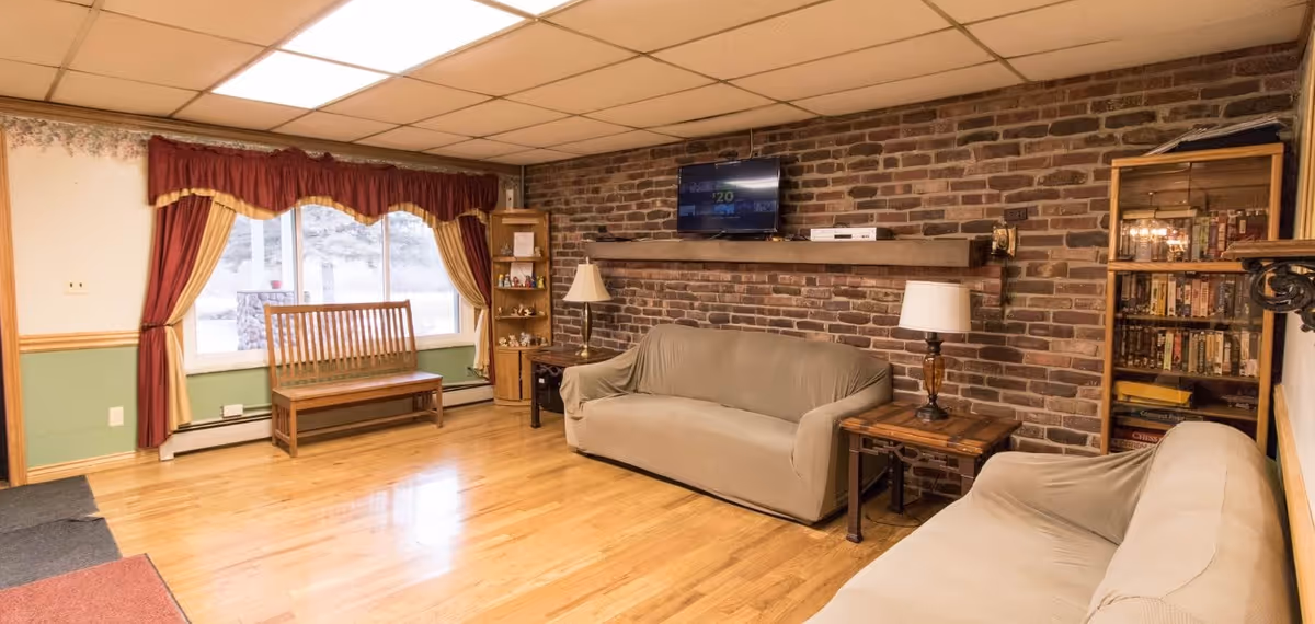 A cozy living room with two beige sofas covered with slipcovers, a wooden bench near a window with red and yellow curtains, a brick accent wall with a mounted flat-screen TV, two wooden side tables with lamps, a wooden shelf with decorative items, and a wooden cabinet filled with books and games.