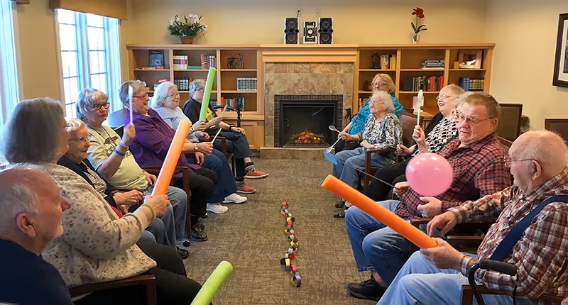 A group of elderly people sitting in two rows facing each other in a cozy room with a fireplace and bookshelves. They are holding colorful foam sticks and some have balloons, appearing to participate in a group activity or game.