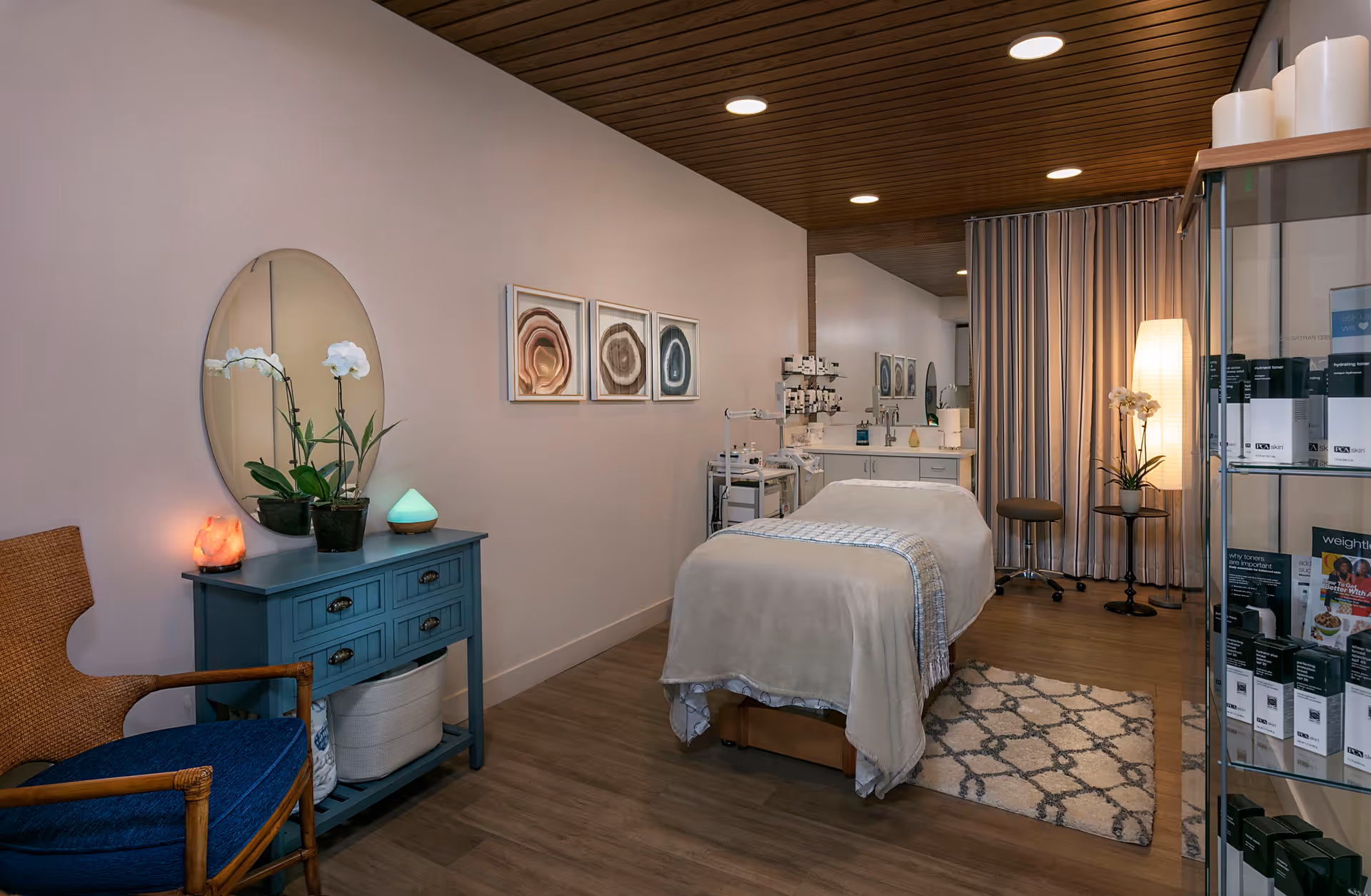 A serene treatment room with a massage table covered in a beige blanket, a blue cabinet with a round mirror and potted orchids, a wicker chair with a blue cushion, and a glass display case with skincare products. The room has wooden flooring, a patterned rug, and soft lighting from ceiling lights and a floor lamp behind a curtain.