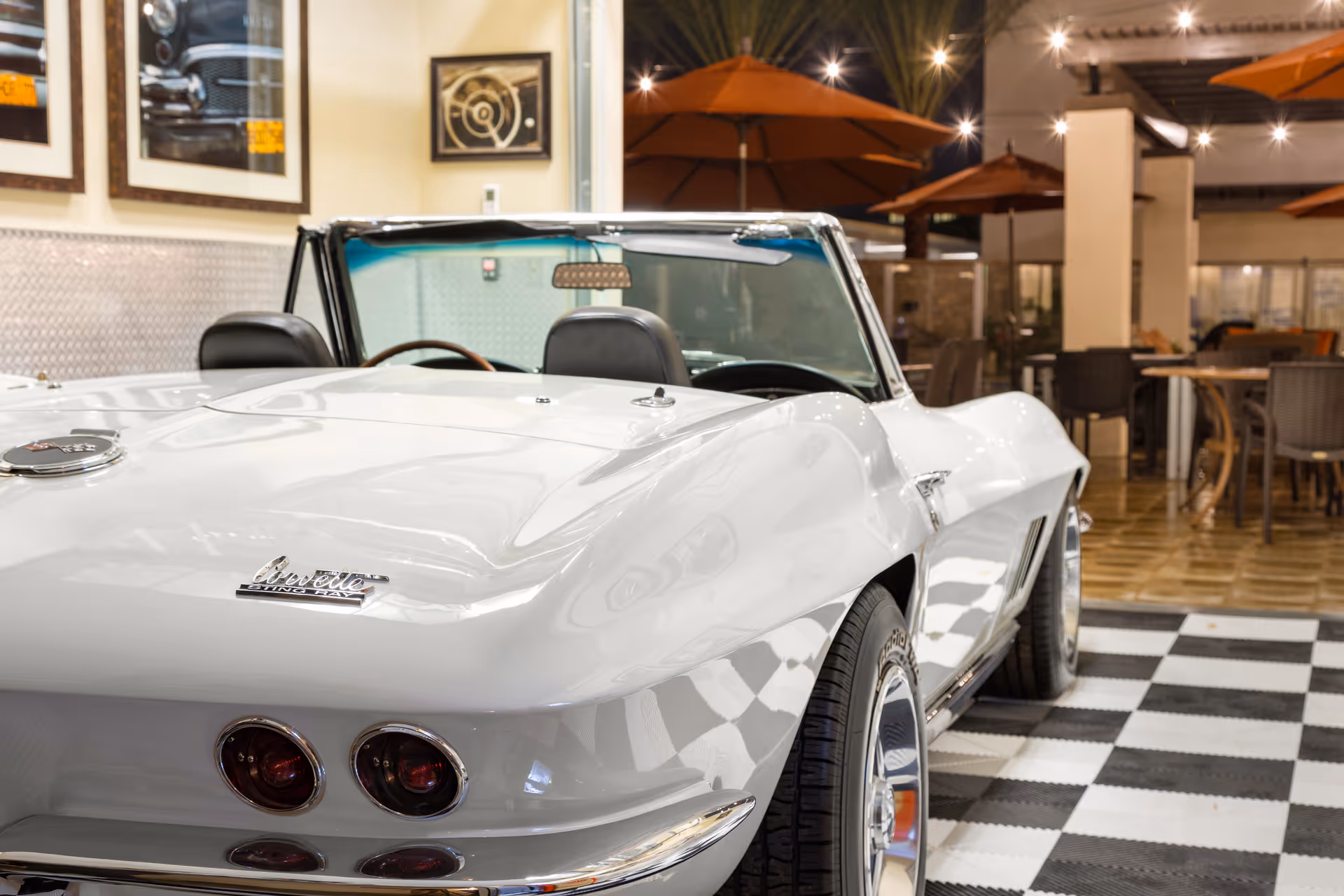 A white classic convertible car displayed indoors on a checkered floor mat, with tables, chairs, and orange umbrellas in the background, suggesting a lounge or common area.