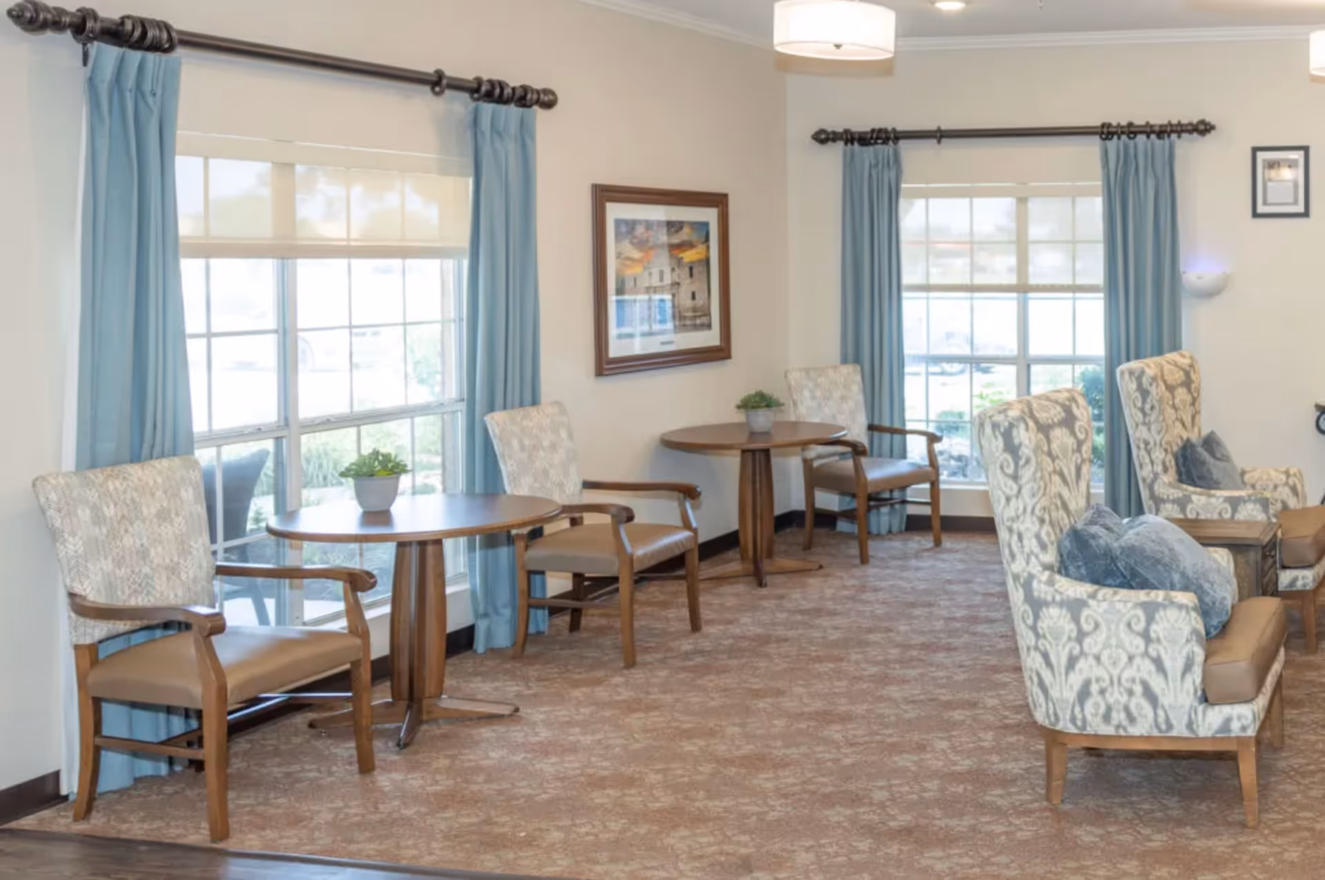 Bright communal sitting area with armchairs, small round tables, large windows with blue curtains and patterned carpet.