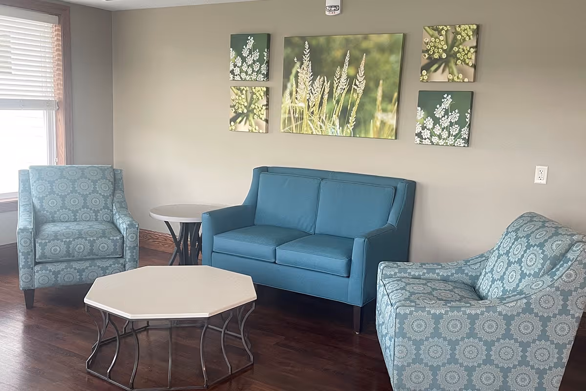 A cozy seating area in a senior living facility featuring a blue loveseat, two patterned blue armchairs, a hexagonal coffee table with a white top and metal legs, and a small round side table. The room has wooden flooring, a window with blinds, and four nature-themed wall art pieces depicting plants and flowers on a beige wall.