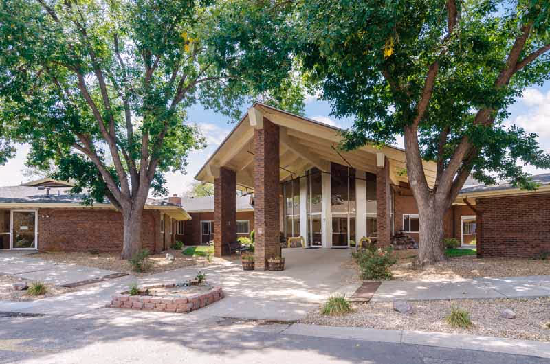 Exterior view of Westlake Lodge Health and Rehabilitation building entrance with large glass windows, brick walls, and a peaked roof supported by brick pillars. There are large trees providing shade and a paved driveway in front.
