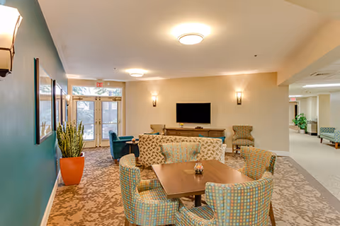 A cozy common area in a senior living facility featuring a wooden table surrounded by patterned upholstered chairs. In the background, there is a wall-mounted flat-screen TV above a wooden console, two armchairs, and a large potted plant near glass double doors letting in natural light. The room has soft lighting from ceiling fixtures and wall sconces, with a carpeted floor and a teal accent wall on the left.