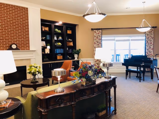 Bright communal living room with seating, a fireplace, built-in bookshelves, piano, and floral arrangements.