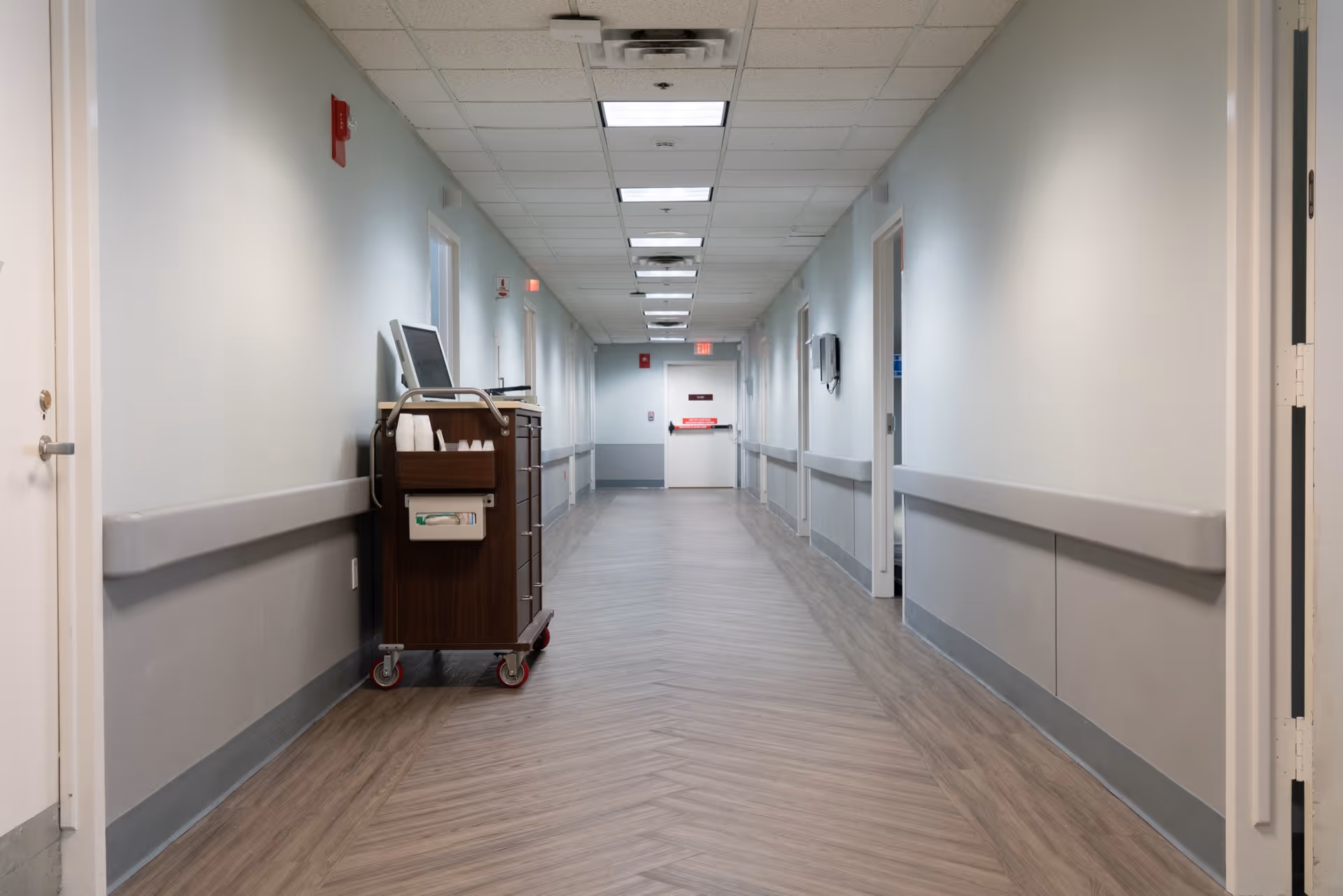 A long, empty hallway in a healthcare facility with light gray walls and wood-patterned flooring. There are handrails along both sides of the hallway, several closed doors, and a medical cart with supplies and a computer on the left side. The ceiling has recessed lighting and ventilation panels.